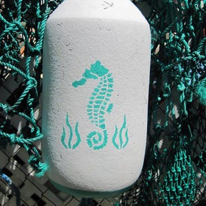 May include: White buoy with a teal blue seahorse and seaweed design painted on the side. The buoy is surrounded by green fishing net.
