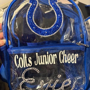 May include: A clear plastic backpack with blue trim and straps. The front features a blue rhinestone horseshoe logo, the white text "Colts Junior Cheer," and the name "Evie" written in cursive script with clear rhinestones.