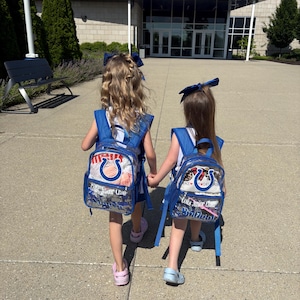 May include: Two clear plastic backpacks with blue trim and straps, each featuring a blue horseshoe logo and the text Colts Junior Cheer. The backpacks are worn by two individuals with long hair, each wearing a large blue hair bow and light-colored shoes.