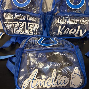 May include: Three clear and blue backpacks with the text "Colts Junior Cheer" and individual names. Each backpack features a blue horseshoe logo and blue straps. The names "Wesley", "Keely", and "Amelia" are displayed in white.