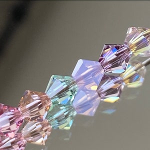 May include: Close-up of a strand of faceted crystal beads in shades of pink, lavender, and aqua. The beads are arranged in a diagonal line, reflecting light and creating a shimmering effect. Ideal for jewelry making.