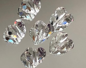 May include: Six clear crystal heart-shaped beads with a faceted design. The beads are scattered on a light gray surface.