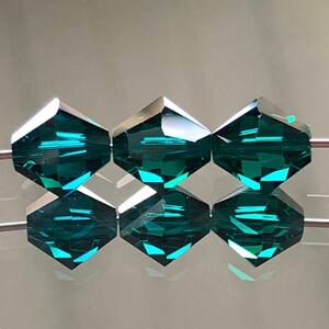 May include: Six teal green faceted glass beads strung on a wire. The beads are hexagonal in shape and have a shiny, reflective surface.