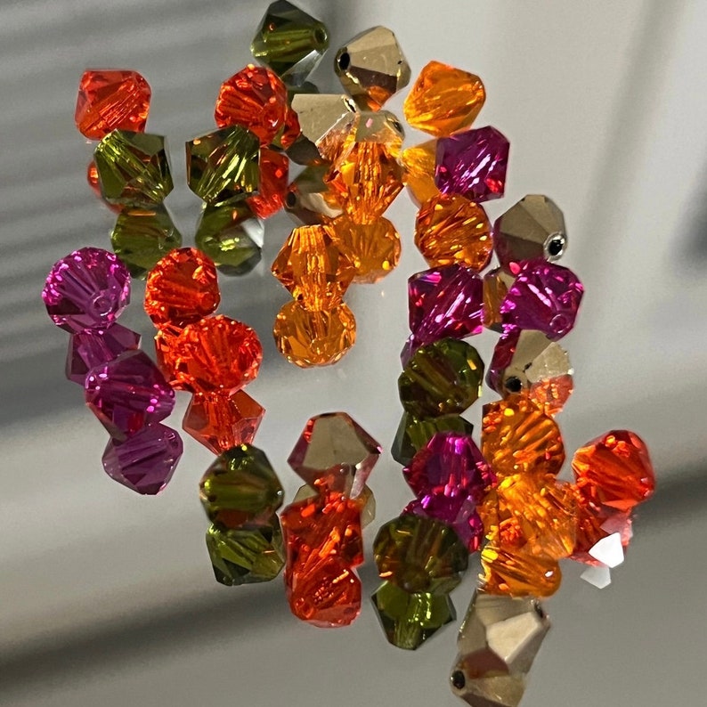 May include: Assortment of faceted glass beads in various colors, including orange, red, pink, and green. The beads are arranged on a reflective surface.