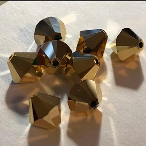 May include: A group of eight gold-colored faceted glass beads. The beads are shaped like cones with a pointed top and a flat bottom. They are arranged in a loose pile on a white surface.