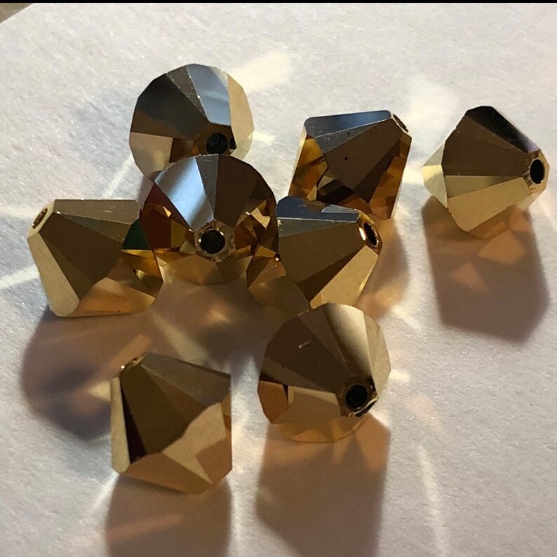 May include: A group of eight gold-colored faceted glass beads. The beads are shaped like cones with a pointed top and a flat bottom. They are arranged in a loose pile on a white surface.