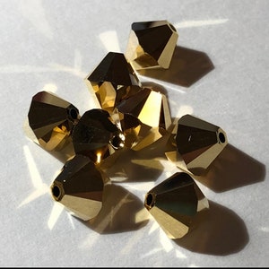 May include: A group of eight gold-colored faceted glass beads. The beads are shaped like cones with a pointed top and a flat bottom. They are arranged in a pile on a white surface.