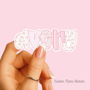 May include: A die-cut sticker featuring the name Avery in lowercase letters. Each letter displays a different pattern in shades of pink and white, including floral prints, gingham checks, and vertical stripes, all outlined in a thick white border.