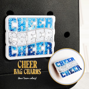May include: Three white and blue glitter "CHEER" charms. The charms are stacked on top of each other. The charms are on a black background. The text "CHEER BAG CHARMS" is written in gold below the charms. The text "Your team colors!" is written in gold below the text "CHEER BAG CHARMS".