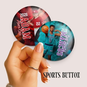 May include: Two sports buttons are held in a hand. One button features a baseball player in a red uniform with the name "Adam" and the text "River Cats". The other button features a basketball player in a teal uniform with the name "Marcus" and the text "East Jaguars". The text "Sports Button" is at the bottom.