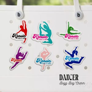 May include: A white tote bag featuring six colorful decorative charms shaped like dancers in various poses. Each charm displays the text Kensie BORN TO DANCE in different colors including red, teal, green, purple, pink, and blue silhouettes.