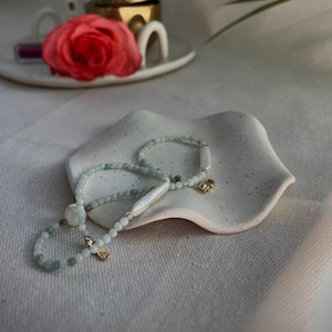 May include: Two delicate bracelets with green jade beads and white pearl accents on a white speckled ceramic dish.