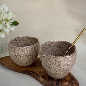 May include: Two speckled ceramic bowls with a gold spoon rest on a wooden serving board. The bowls have a light brown base color with dark brown speckles. White flowers are in the background.