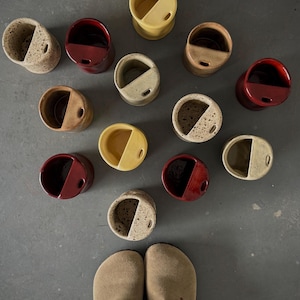 Peut inclure: Une collection de onze tasses en céramique disposées en groupe, chacune dotée d'une étagère interne semi-circulaire et d'un petit trou sur le bord. Les tasses présentent diverses finitions, dont beige moucheté, jaune uni, rouge foncé et marron.