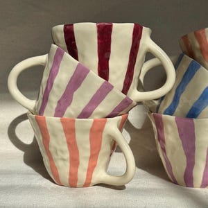 May include: A collection of handmade ceramic mugs with vertical stripes in various colors, including red, purple, pink, and blue. The mugs have a textured surface and a curved handle. The mugs are stacked together, showcasing their unique design.