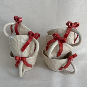 May include: A set of six off-white ceramic teacups with textured surfaces. Each cup is adorned with a bright red bow on the handle. The cups are stacked in pairs, creating a charming and festive display.