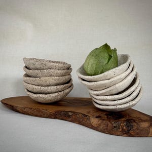 May include: A stack of small, speckled ceramic bowls in a natural color, next to a stack of white speckled bowls, one holding a green tomatillo. The bowls are on a wooden serving board.