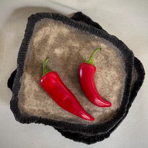 May include: Two vibrant red chili peppers rest on a textured, beige tray with a dark, fringed edge. The peppers have green stems and a glossy appearance, contrasting with the tray's earthy tones. The tray is set against a neutral background.