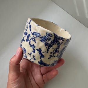 May include: A cream-colored ceramic cup with an irregular, hand-formed shape. The exterior features a detailed blue ink-style pattern depicting various birds, dragonflies, and floral branches scattered across the textured, dimpled surface.