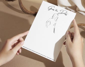 May include: A white card with the text "Guess the Dress" written in black cursive. A simple line drawing of a woman in a dress is in the center of the card.