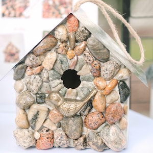 May include: A decorative birdhouse covered in various sized, colorful stones. A small, gold-colored bird figurine is perched below the entry hole. The birdhouse has a rope loop for hanging.
