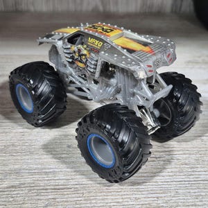 May include: A silver and yellow MAX-D monster truck toy with large black tyres and blue accents. The truck features a skull graphic on the side and the name "MAX-D" on the roof.