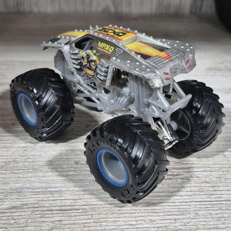 May include: A silver and yellow MAX-D monster truck toy with large black tyres and blue accents. The truck features a skull graphic on the side and the name "MAX-D" on the roof.