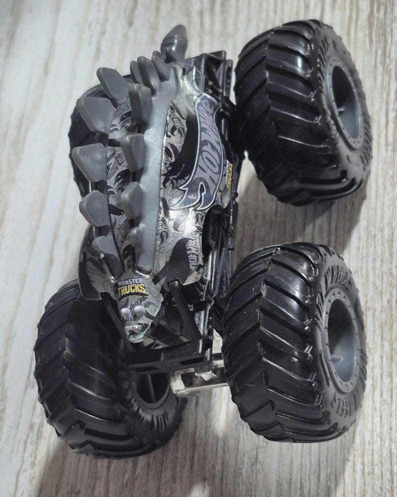 May include: A black and silver Hot Wheels Monster Truck with large, textured tyres. The truck features a detailed design with the Hot Wheels logo and the words "Monster Trucks". The truck has a spiked design on top.
