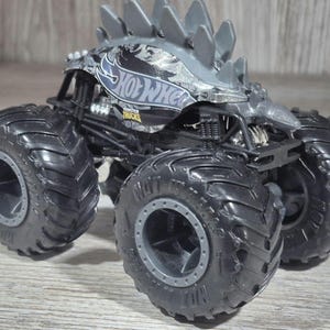 May include: A black and grey Hot Wheels monster truck toy with large tyres. The truck has a grey spiked roof and the Hot Wheels logo on the side. The tyres are black with grey rims.