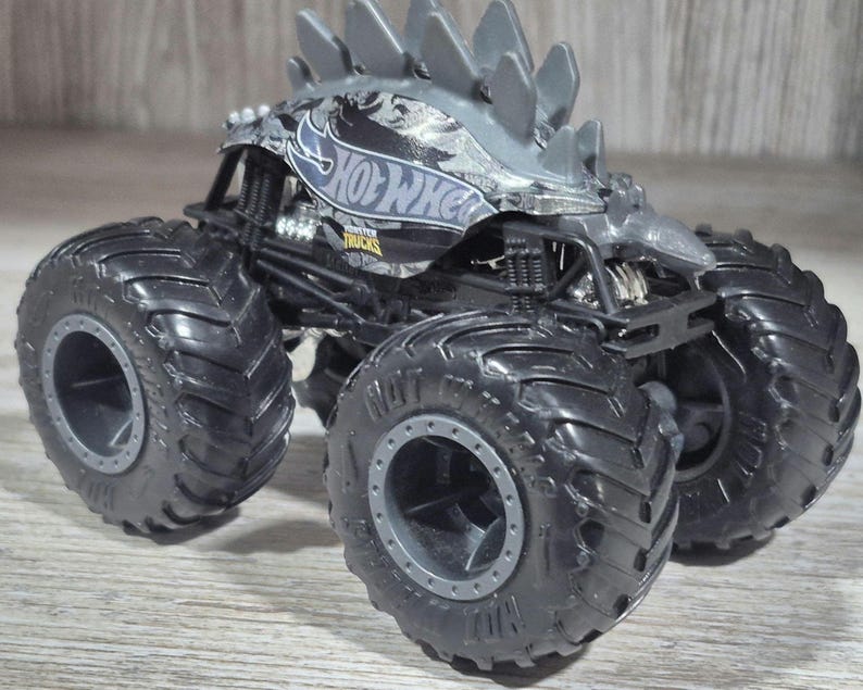 May include: A black and grey Hot Wheels monster truck toy with large tyres. The truck has a grey spiked roof and the Hot Wheels logo on the side. The tyres are black with grey rims.