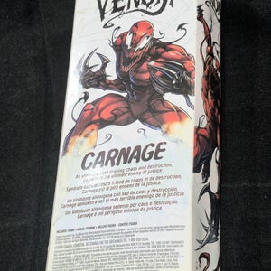May include: A rectangular white cardboard packaging box for a Marvel Venom Carnage action figure. The front features a large, detailed illustration of the red and black Carnage character with sharp claws, the Venom logo, and descriptive text.