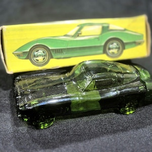 May include: A translucent green glass car figurine shaped like a sports car, positioned in front of its original rectangular yellow cardboard box. The box features a printed illustration of a green sports car on its front panel.