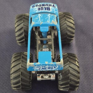 May include: A blue toy monster truck with large black tires. The truck has the words "BLUE THUNDER" and "FEEL THE ENERGY" printed on it. Lightning bolt graphics are visible on the truck's body.