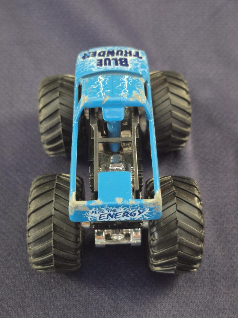 May include: A blue toy monster truck with large black tires. The truck has the words "BLUE THUNDER" and "FEEL THE ENERGY" printed on it. Lightning bolt graphics are visible on the truck's body.