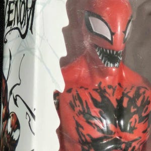 May include: A red and black Carnage action figure from the Marvel Venom series, encased in a clear plastic box. The packaging features the Marvel Venom logo and a graphic illustration of the character on the side panel, with white web patterns.