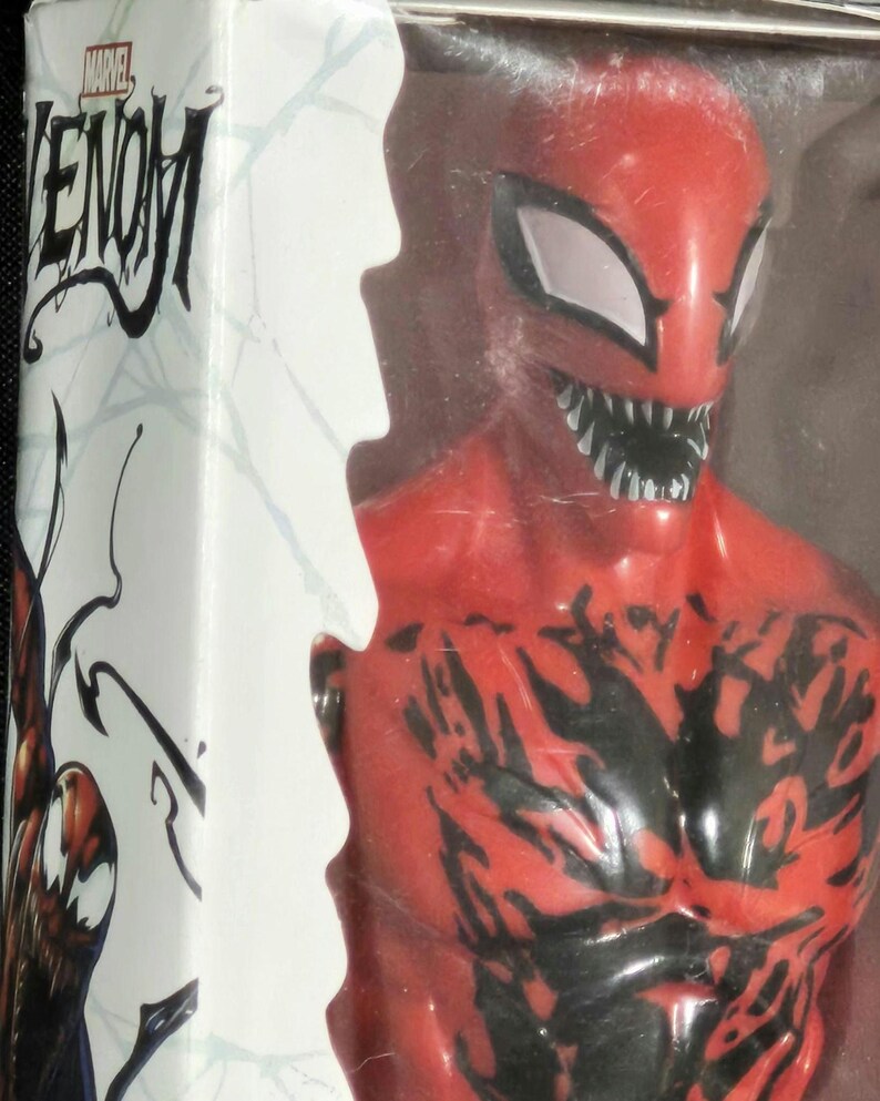 May include: A red and black Carnage action figure from the Marvel Venom series, encased in a clear plastic box. The packaging features the Marvel Venom logo and a graphic illustration of the character on the side panel, with white web patterns.