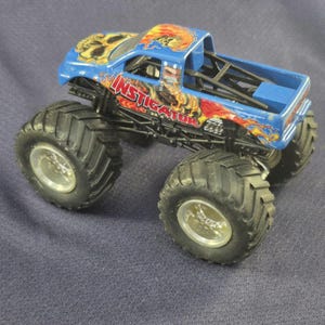 May include: A blue monster truck toy with large black tires and silver rims. The truck features a skull graphic on the hood and the word "INSTIGATOR" in red and orange flames on the side. The truck bed is blue.