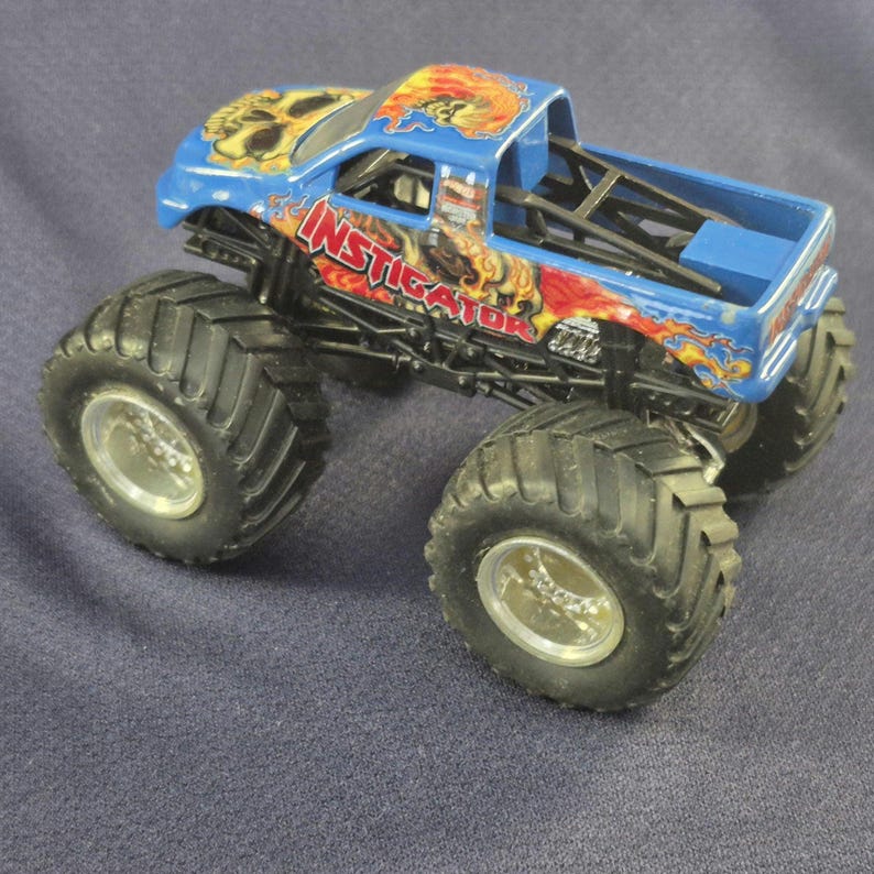 May include: A blue monster truck toy with large black tires and silver rims. The truck features a skull graphic on the hood and the word "INSTIGATOR" in red and orange flames on the side. The truck bed is blue.