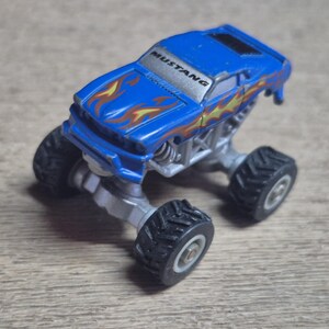 A blue toy monster truck with the word "MUSTANG" on the windshield. The truck has flame decals and large black tires. The chassis is silver.