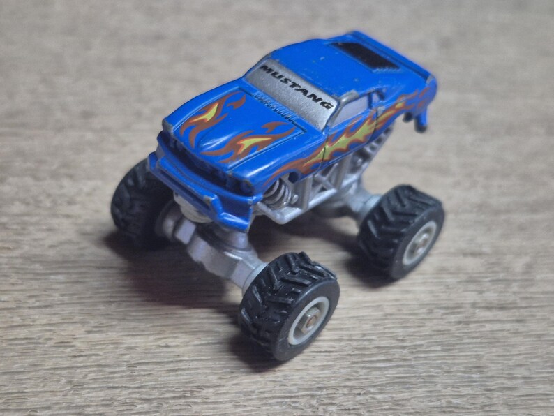 A blue toy monster truck with the word "MUSTANG" on the windshield. The truck has flame decals and large black tires. The chassis is silver.