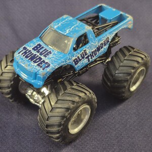 May include: A blue toy monster truck with the words "BLUE THUNDER" printed on the cab. The truck has large black tires and a silver chassis. The truck is a toy vehicle.