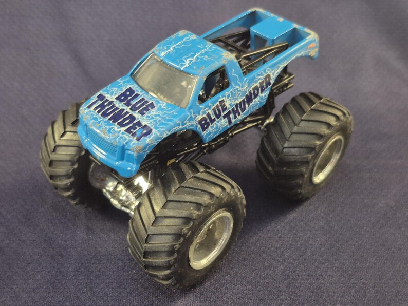 May include: A blue toy monster truck with the words "BLUE THUNDER" printed on the cab. The truck has large black tires and a silver chassis. The truck is a toy vehicle.