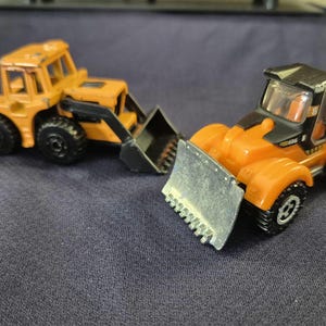 May include: Two toy construction vehicles. One is a yellow and black front-end loader with a silver blade. The other is a yellow bulldozer with a black bucket. Both have black wheels and are on a dark blue surface.