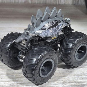 May include: A grey and black Hot Wheels monster truck toy with large black tyres. The truck has a spiked design on top and the Hot Wheels logo on the side.
