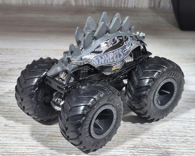 May include: A grey and black Hot Wheels monster truck toy with large black tyres. The truck has a spiked design on top and the Hot Wheels logo on the side.