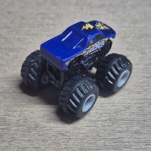A blue and black toy monster truck with large black tires and gray rims. The truck has the word "SHOCKER" in yellow and white lettering on the side. The top of the truck is blue with yellow lightning bolt graphics.