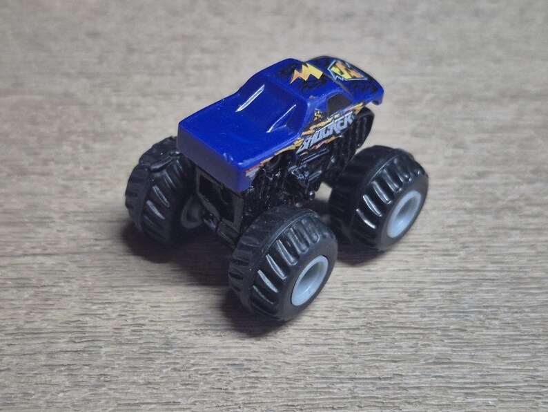 A blue and black toy monster truck with large black tires and gray rims. The truck has the word "SHOCKER" in yellow and white lettering on the side. The top of the truck is blue with yellow lightning bolt graphics.