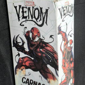 May include: A white rectangular cardboard box featuring Marvel Venom branding and a large illustration of the red and black Carnage character. The box displays the name Carnage in bold black text and includes descriptive text about the character.