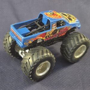 May include: A blue toy monster truck with large black tires and silver rims. The truck has the word "INVEIGATOR" printed on the side in red and yellow flames. The truck's cab has a skull design on top.