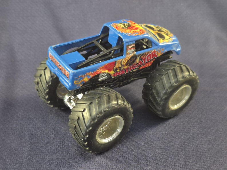 May include: A blue toy monster truck with large black tires and silver rims. The truck has the word "INVEIGATOR" printed on the side in red and yellow flames. The truck's cab has a skull design on top.